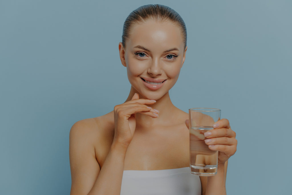 Young cheerful attractive female with clean glowing face skin drinking water to prevent dehydration, smiling charming woman with glass of aqua. Health and nutrition concept